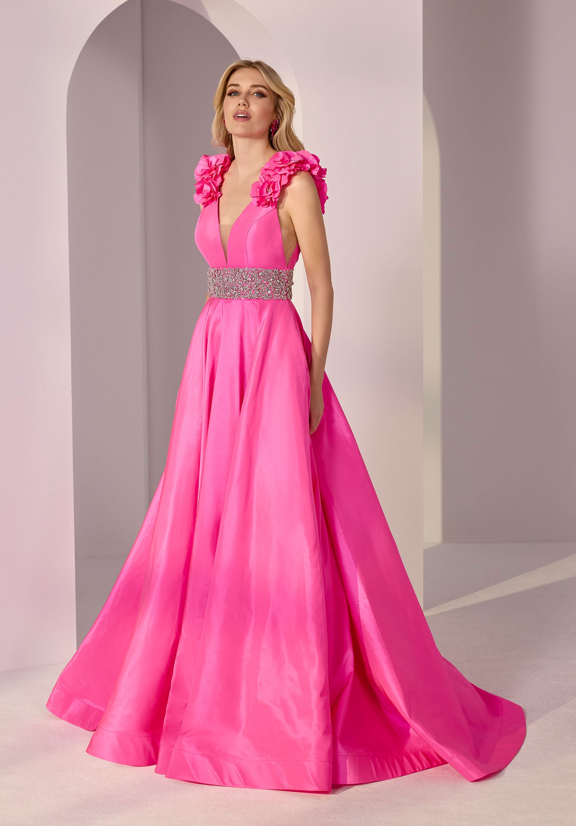 A woman stands in a bright pink Morilee 49202 long prom dress with 3D rosettes on the shoulders and a beaded waist, set against a pale modern backdrop with archways.